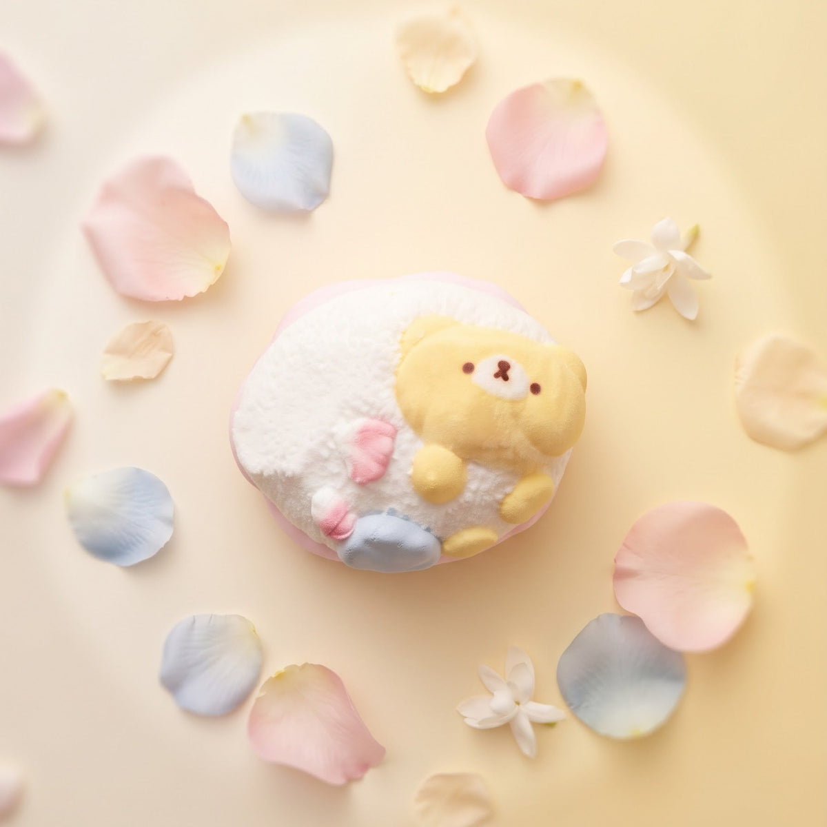 A cute, round white bath bomb or soap featuring a yellow bear character on top, styled in a flat-lay with pastel pink, blue, and cream flower petals on a soft cream-colored background.