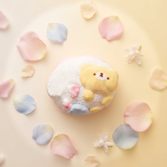 A cute, round white bath bomb or soap featuring a yellow bear character on top, styled in a flat-lay with pastel pink, blue, and cream flower petals on a soft cream-colored background.