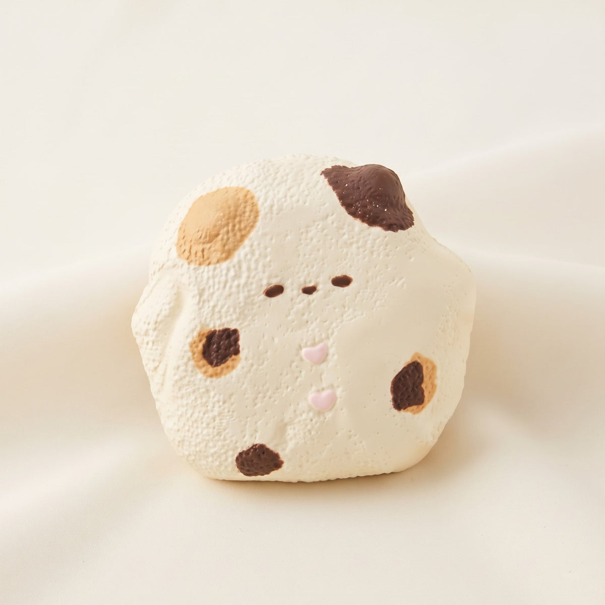 A cute, rounded white plush toy or cushion with a hand-drawn face and spots in dark brown, tan, and light pink, set against a soft cream-colored fabric background.
