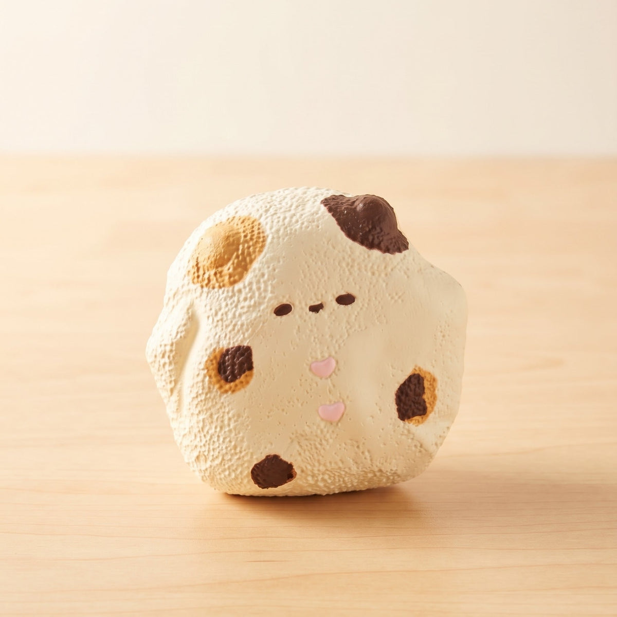 A cute, round, textured squishy toy shaped like a cream-colored cookie with brown spots and a small pink heart on its nose, sitting on a light wooden surface against a warm beige background.