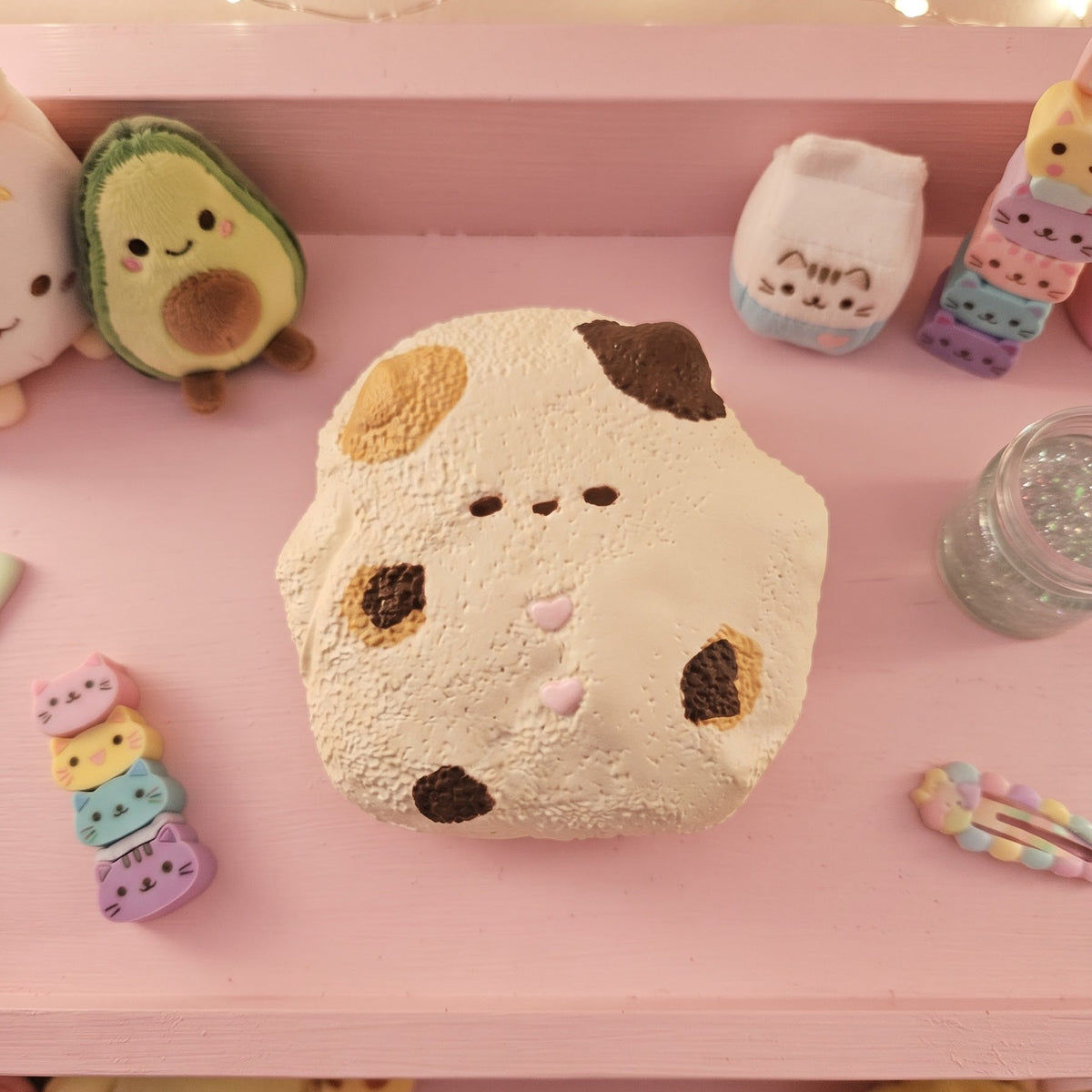 A cute, round, cream-colored plush toy with a cow-like face pattern, displayed on a pink shelf alongside other pastel-colored plush toys and accessories.