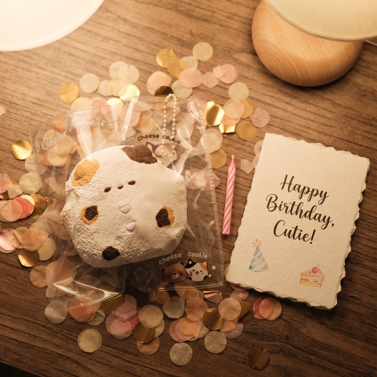 A cute, round, cat-shaped plush toy with a white body, brown ears, and small embroidered facial features, packaged in clear cellophane and placed on a wooden table surrounded by gold and pink confetti. Next to it is a small card that reads 'Happy Birthday, Cute!' and a pink striped candlestick.