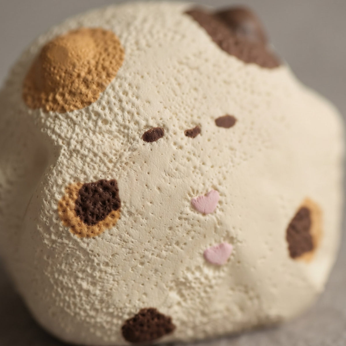 A close-up of a small, rounded, textured cream-colored plush toy or figurine with brown spots and pink nose details, photographed on a soft gray surface.