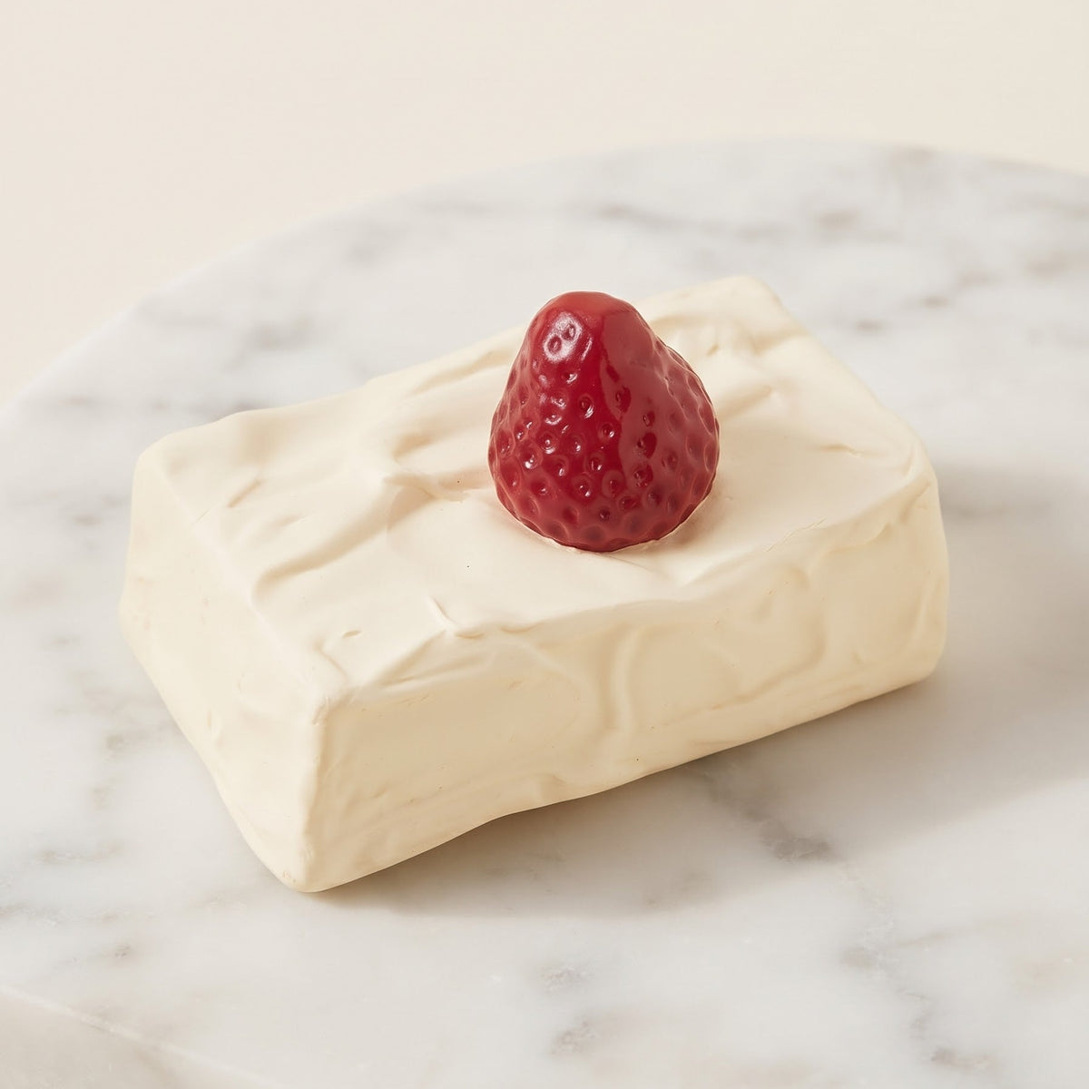A rectangular cream-colored cheese block with a textured rind, topped with a small red strawberry, presented on a white marble serving board against a warm cream background.