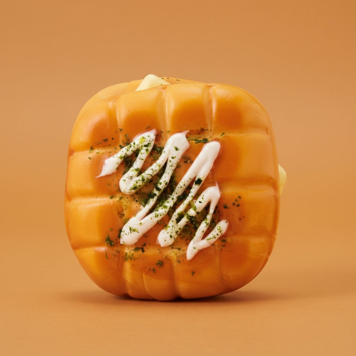 A pumpkin-shaped pastry or bread with a golden-orange baked surface, topped with white drizzle and green herb flakes, centered on a warm tan background.