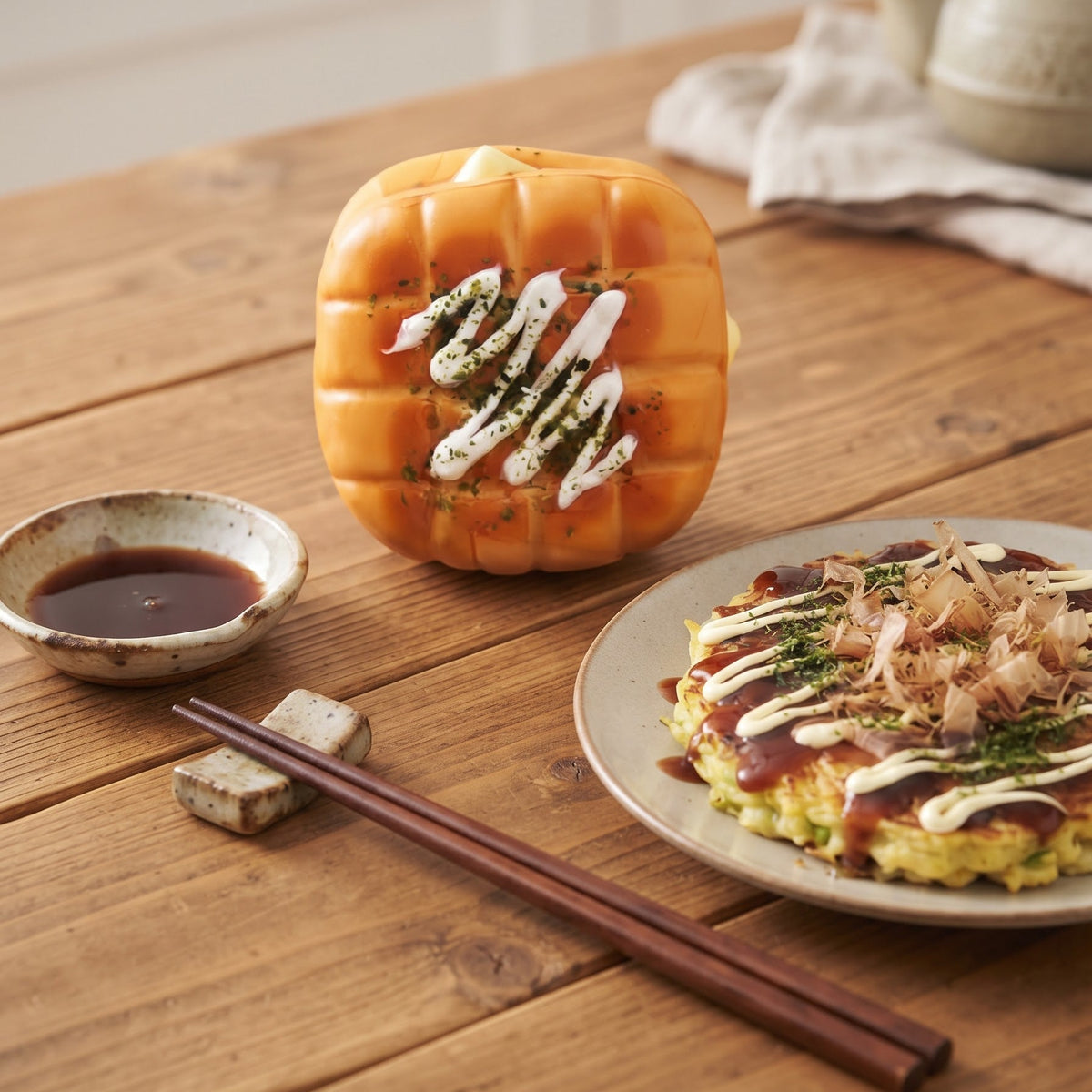 A Japanese-style octagonal bread or pastry topped with white sauce, green herbs, and red sauce sits on a wooden table beside a plate of similar food, a small bowl of dipping sauce, chopsticks, and a teapot in a softly blurred kitchen setting.