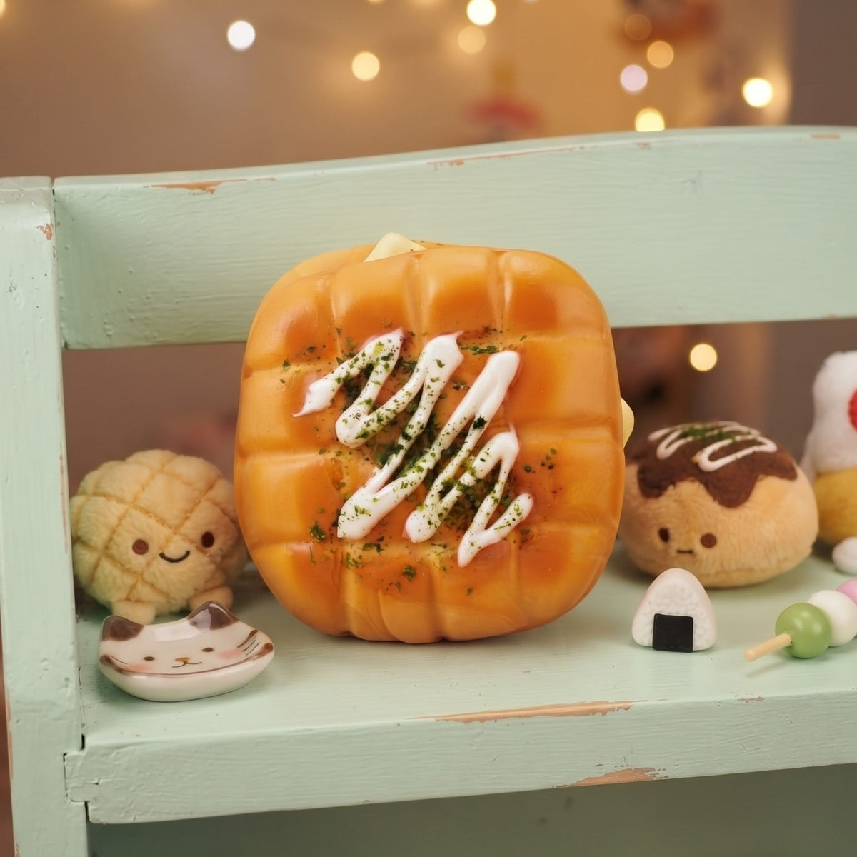 A cute, pumpkin-shaped squishy toy designed to look like a toasted bread roll with white drizzle and green herb garnish, displayed on a light green wooden shelf alongside other small plush food toys.