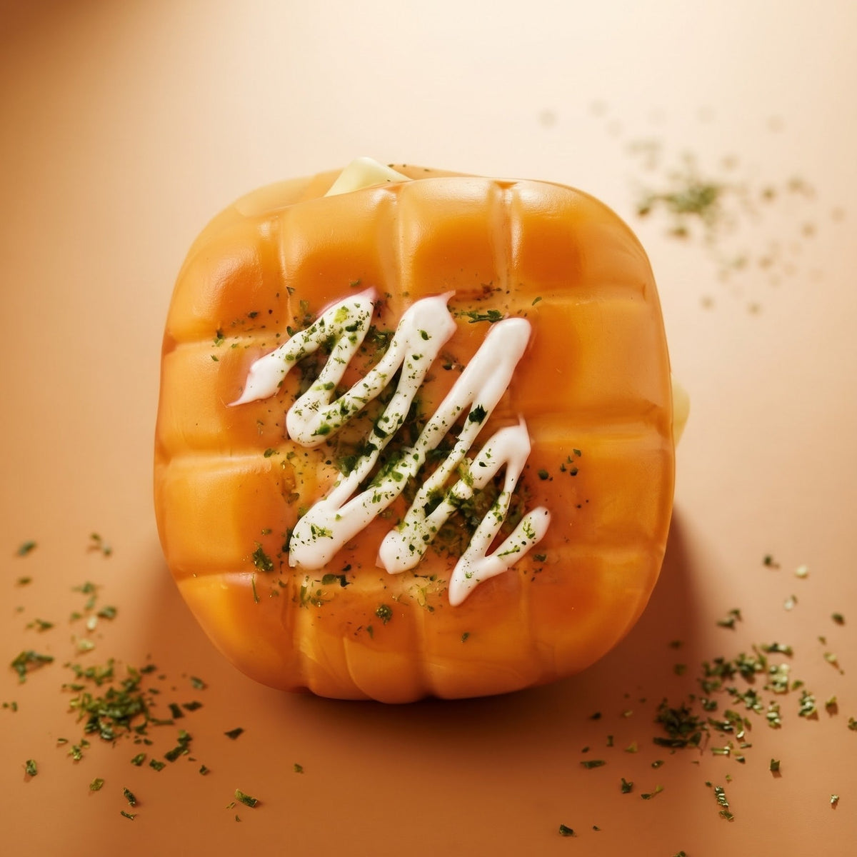 A pumpkin-shaped baked good or pastry topped with white drizzle and green herb garnish, photographed on a warm beige background with scattered herb crumbs.
