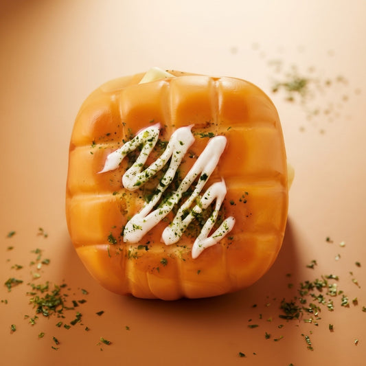 A pumpkin-shaped baked good or pastry topped with white drizzle and green herb garnish, photographed on a warm beige background with scattered herb crumbs.