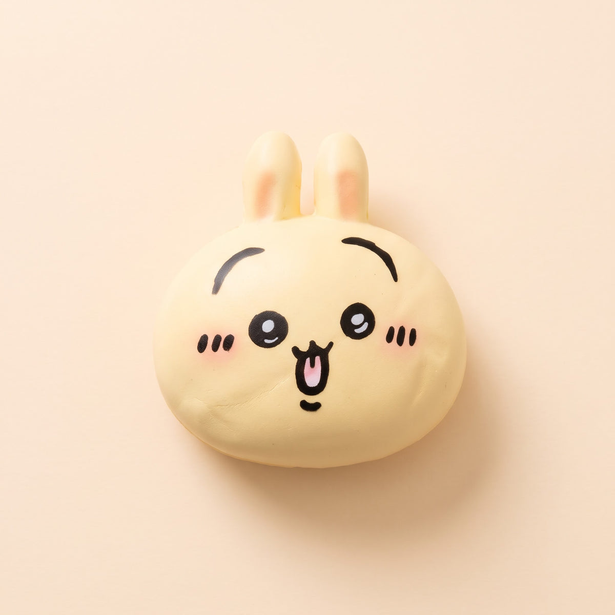 A cute, round, pale yellow bunny-shaped squishy toy with black line art eyes, nose, and mouth, and pink blush marks on its cheeks, set against a solid pale peach background.
