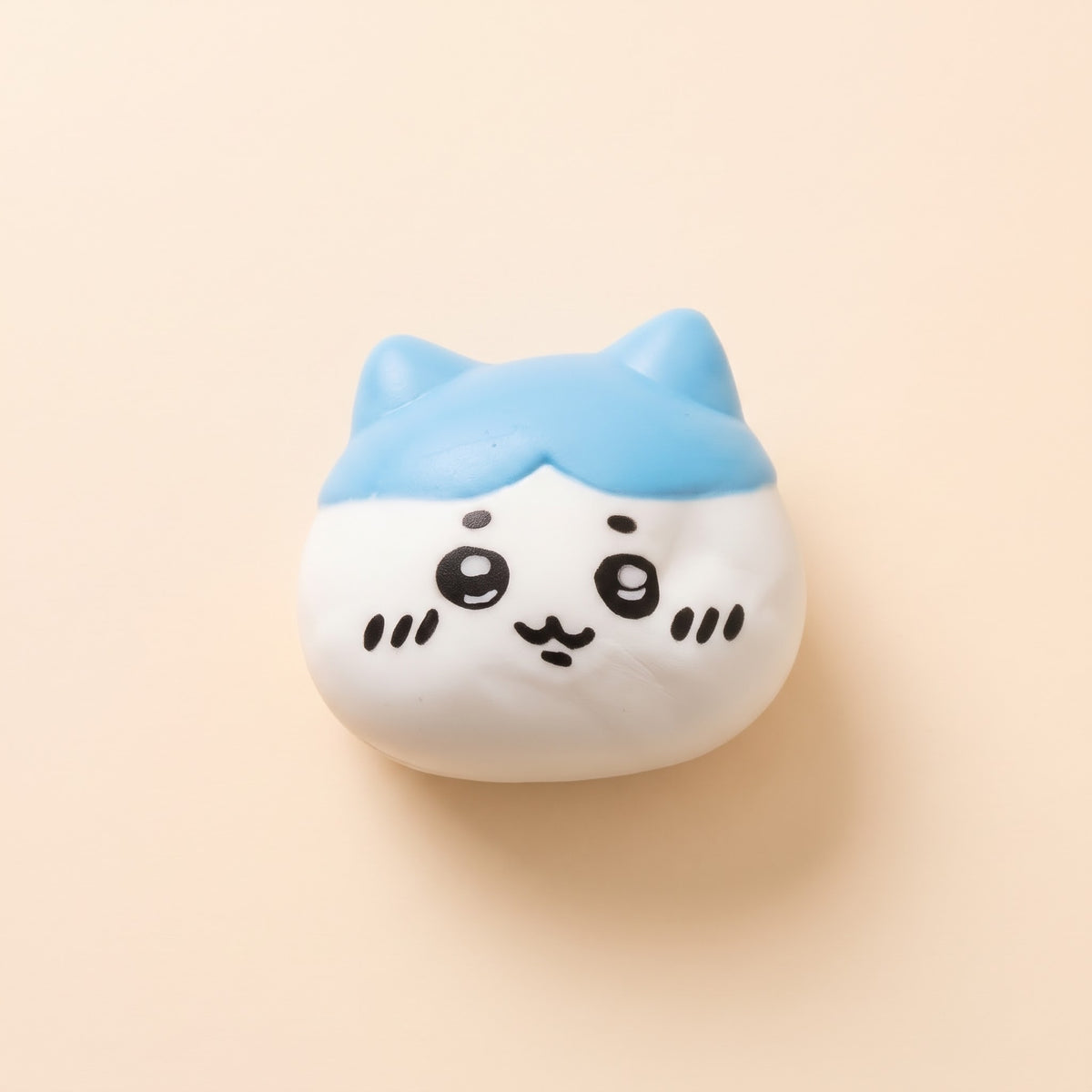 A cute, round white character toy with a light blue cat-ear top and simple black facial features, photographed on a solid pale peach background.