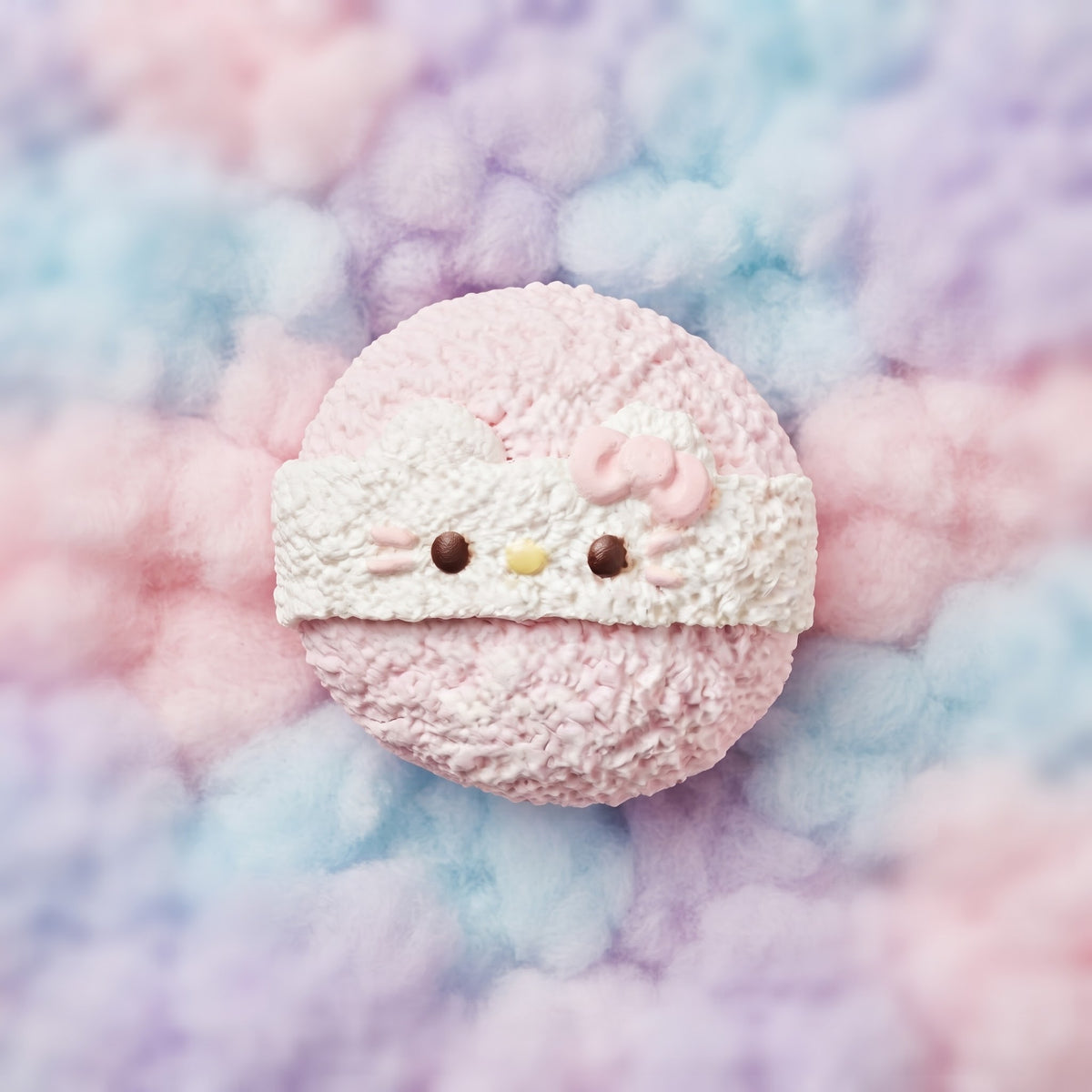A round pink macaron decorated to look like Hello Kitty, featuring a white textured bandana, pink bow, and small yellow nose, set against a soft pastel pink, blue, and purple cloud-like background.