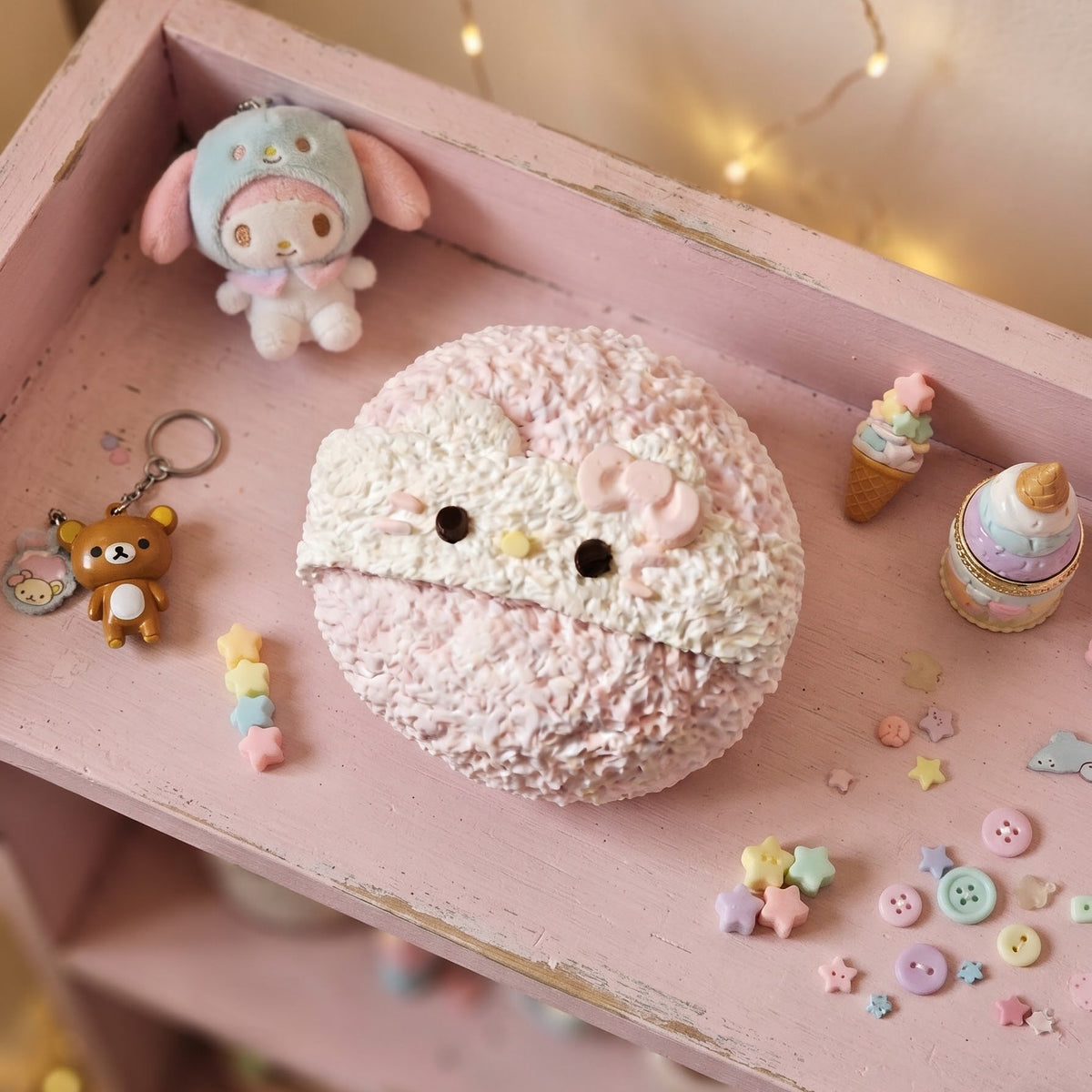 A pastel pink textured round plush toy or cushion with a cute face, displayed on a distressed pink wooden shelf alongside small decorative items and string lights.
