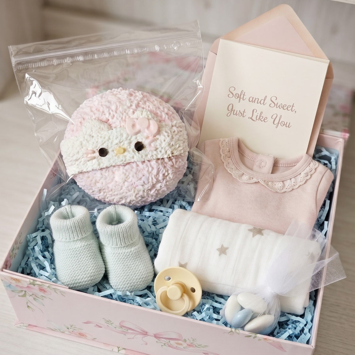 A baby gift set arranged in a pink floral box with blue shredded paper. The set includes a pink plush toy, a pale pink baby outfit, light blue knit booties, a white blanket with star details, a pacifier, and a card that reads 'Soft and Sweet, Just Like You.