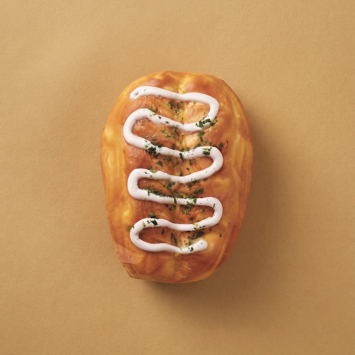 A single baked bread roll shaped like a football, topped with white drizzle and chopped green herbs, centered on a warm tan background.