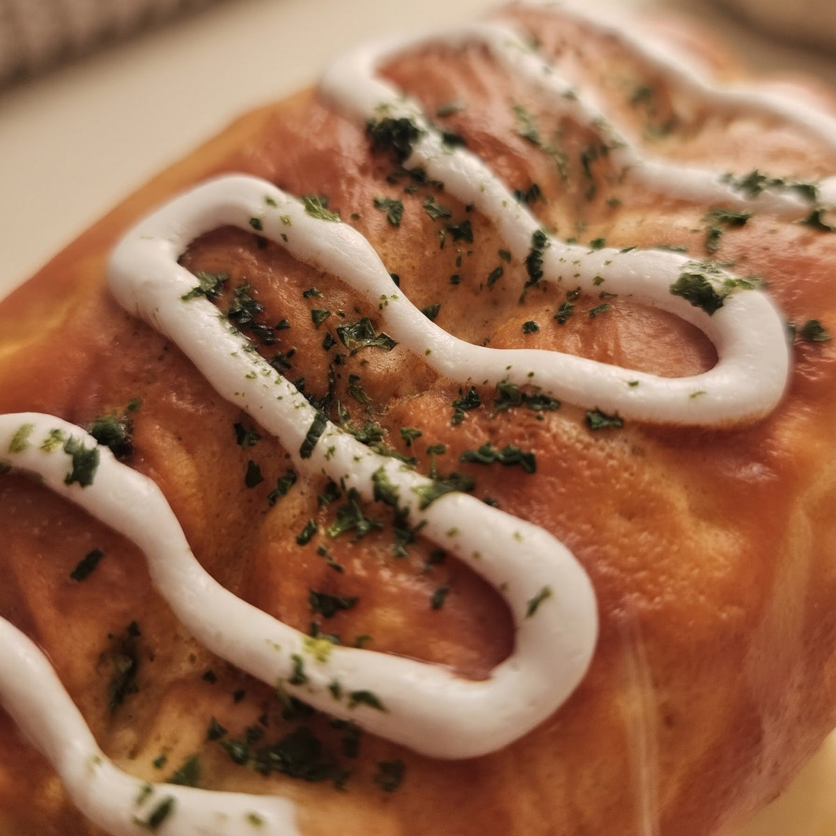 A close-up shot of a golden-brown baked pastry, likely a cinnamon roll or bread roll, decorated with a zigzag drizzle of white icing and sprinkled with chopped green herbs, set against a warm, softly blurred background.