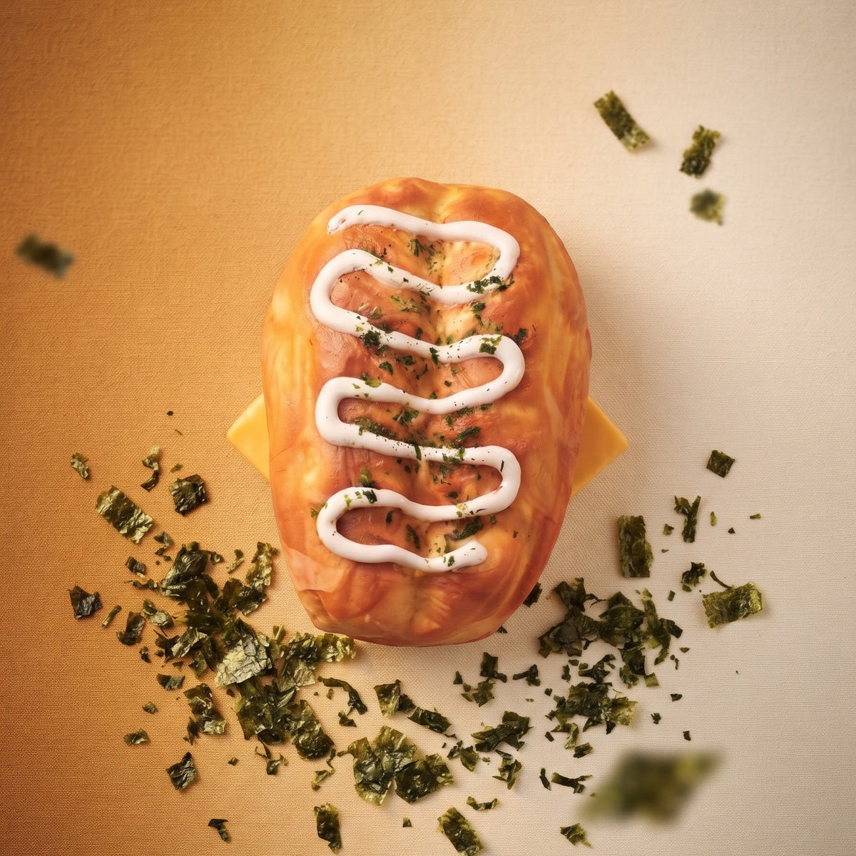 A football-shaped pastry or bread with a glossy orange-brown glaze, white drizzle, and green herb topping, photographed from above on a warm beige background with scattered green herb pieces around it.