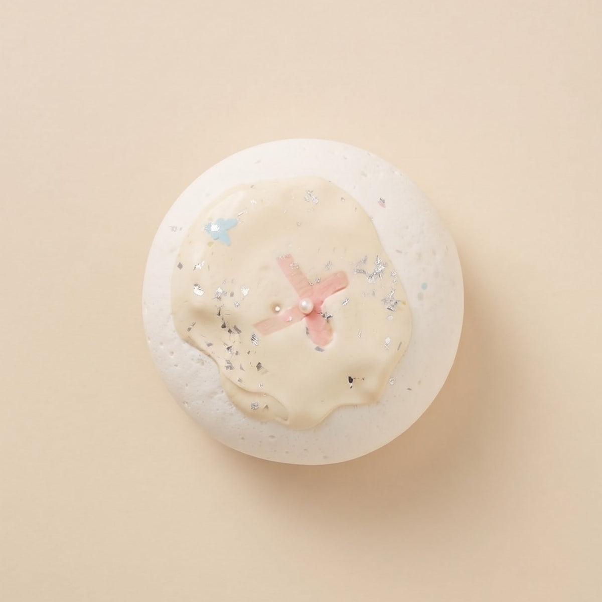 A round white bath bomb with a cream-colored center, a pink cross motif, and small blue and silver glitter accents, photographed from directly above on a solid beige background.