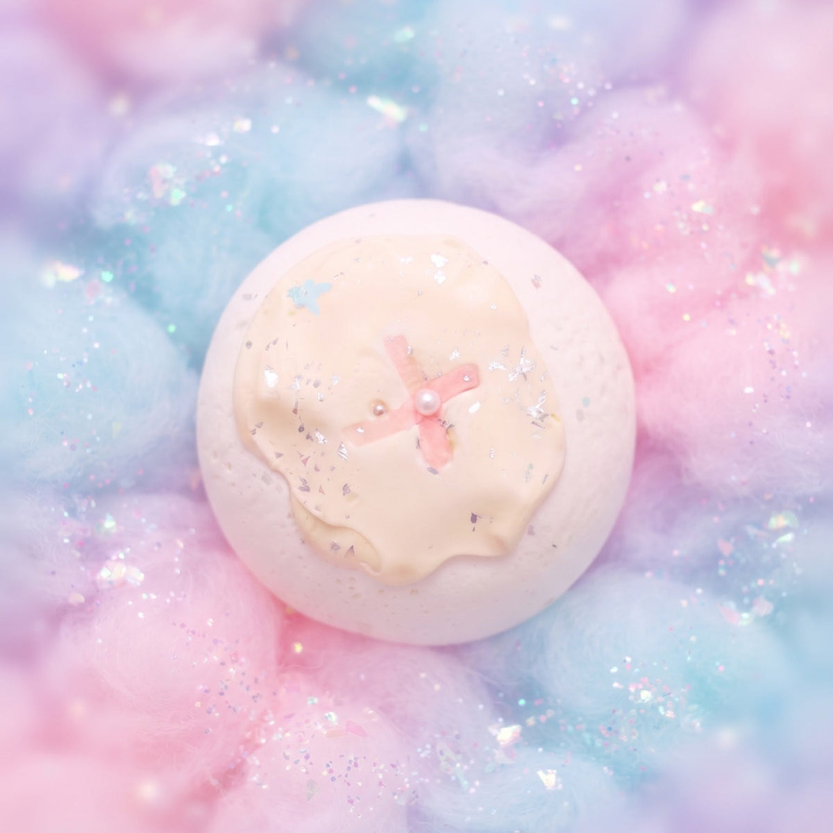 A round, pale pink bath bomb or soap resting on a bed of fluffy pastel pink and blue cotton candy with silver glitter.