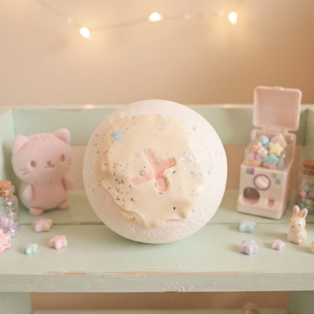 A round bath bomb with a textured, speckled white surface and a pink cross design in the center, displayed on a pastel green shelf with cute decorative accessories and warm fairy lights in the background.