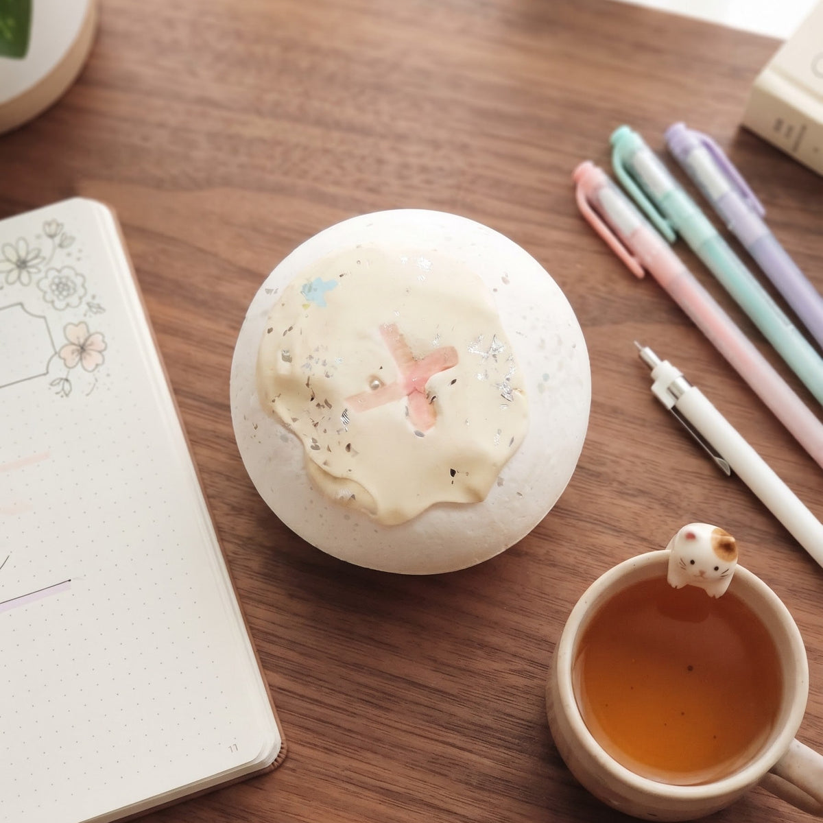 A round white ceramic container with a textured cream-colored lid featuring a small pink airplane and blue speckles, styled on a wooden desk next to a notebook, pens, and a cup of tea.