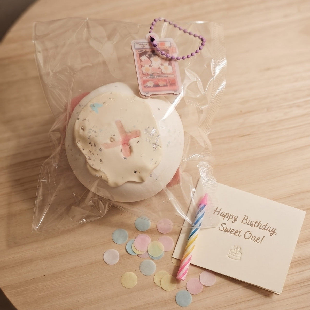 A round white cake-shaped squishy toy with pink frosting details, shown inside a clear plastic bag on a light wood table, alongside a small birthday card that reads 'Happy Birthday Sweet One!' and scattered pastel confetti.