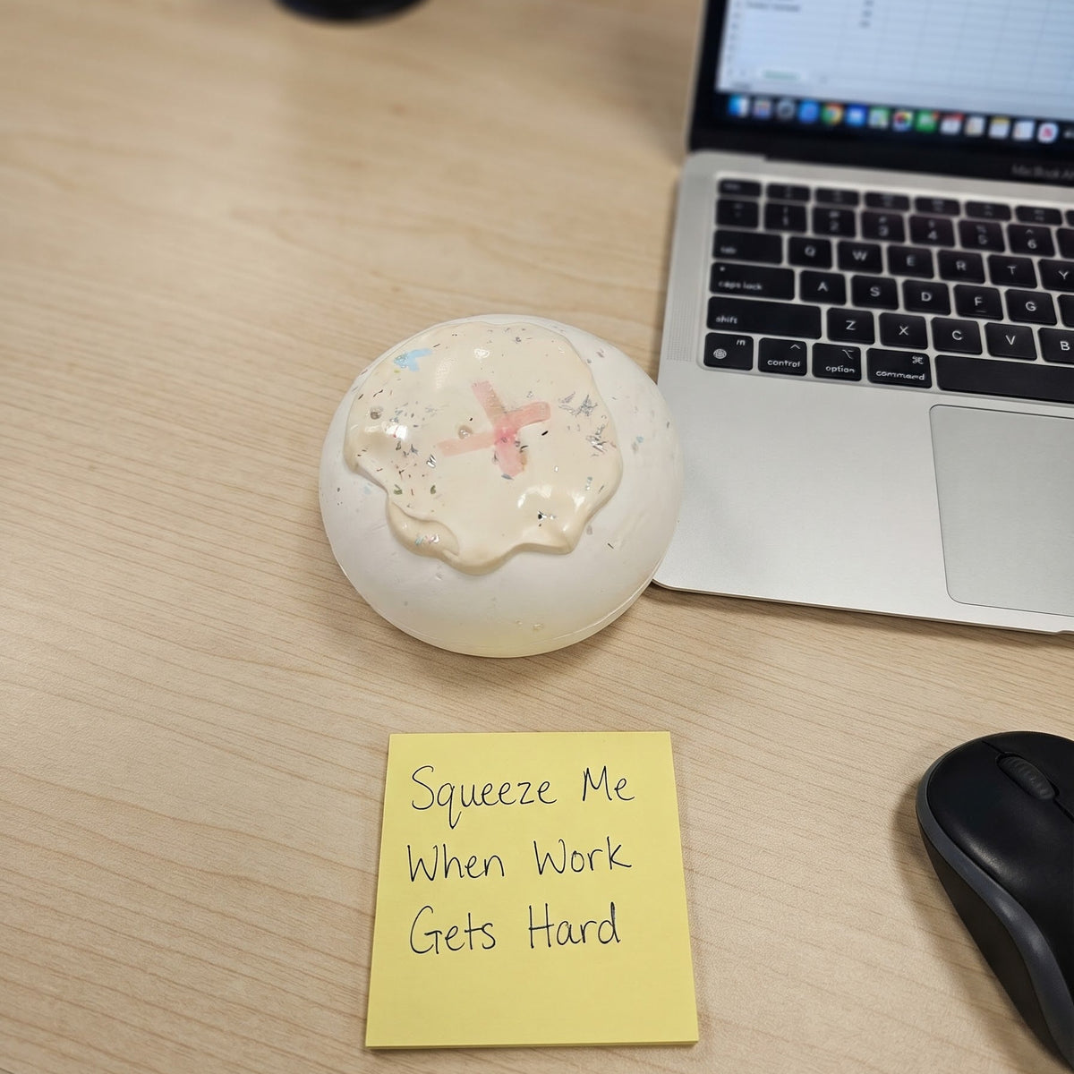 A round, off-white decorative object with a small pink butterfly and speckled pattern sits on a light wood desk next to a laptop, a black computer mouse, and a yellow sticky note that reads 'Squeeze Me When Work Gets Hard'.