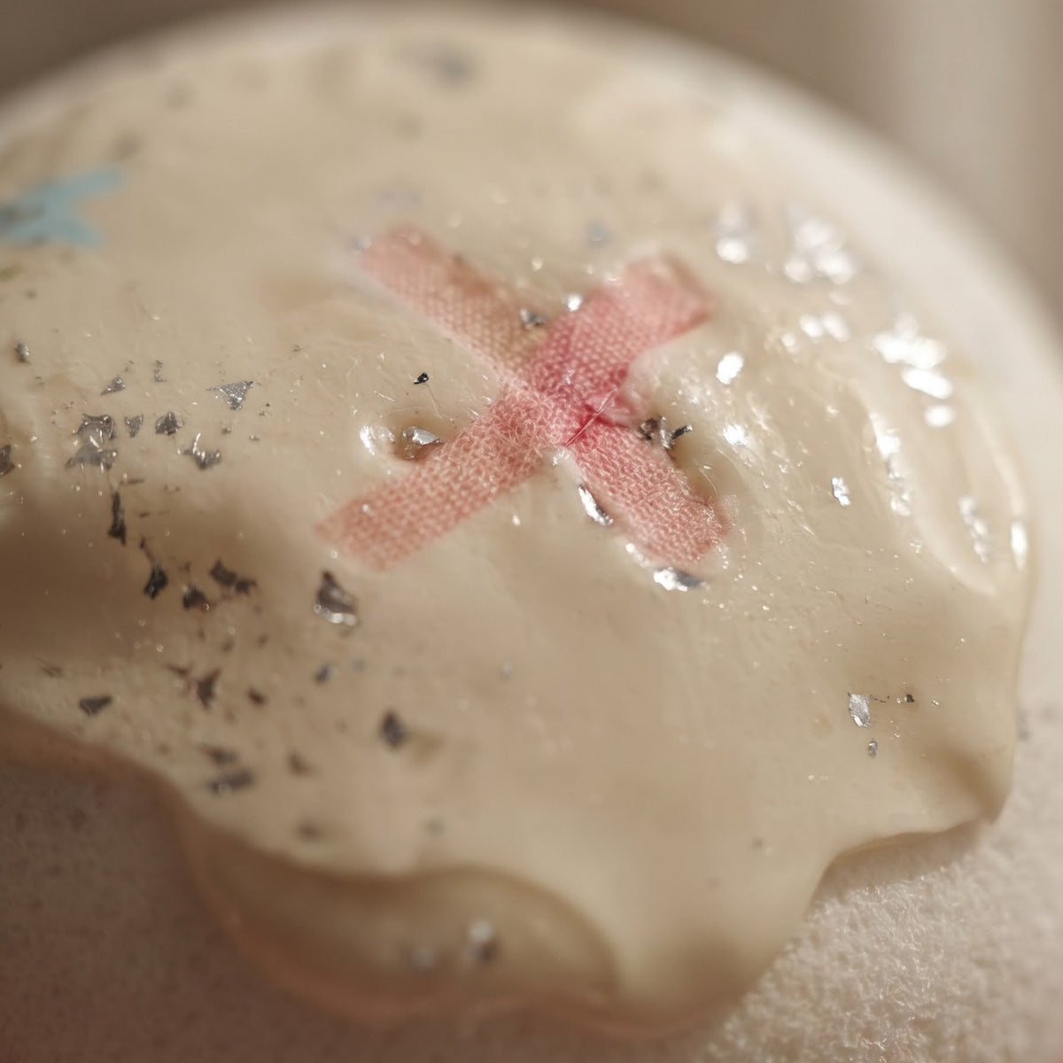 A close-up of a round bath bomb with a beige, cream-colored surface, featuring a pink stitched 'X' on a bandage design, scattered black glitter, and small blue star accents.