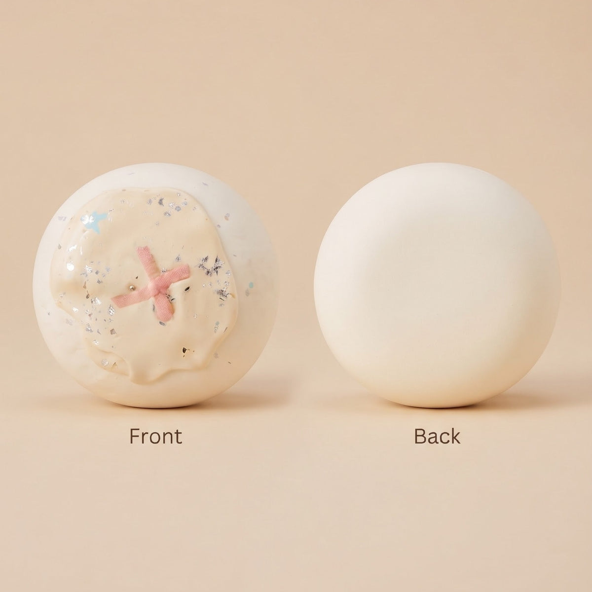 Two round cream-colored bath bombs labeled 'Front' and 'Back' on a warm beige background. The left 'Front' bath bomb shows a textured, cracked surface with a pink cross in the center and tiny blue glitter flecks.