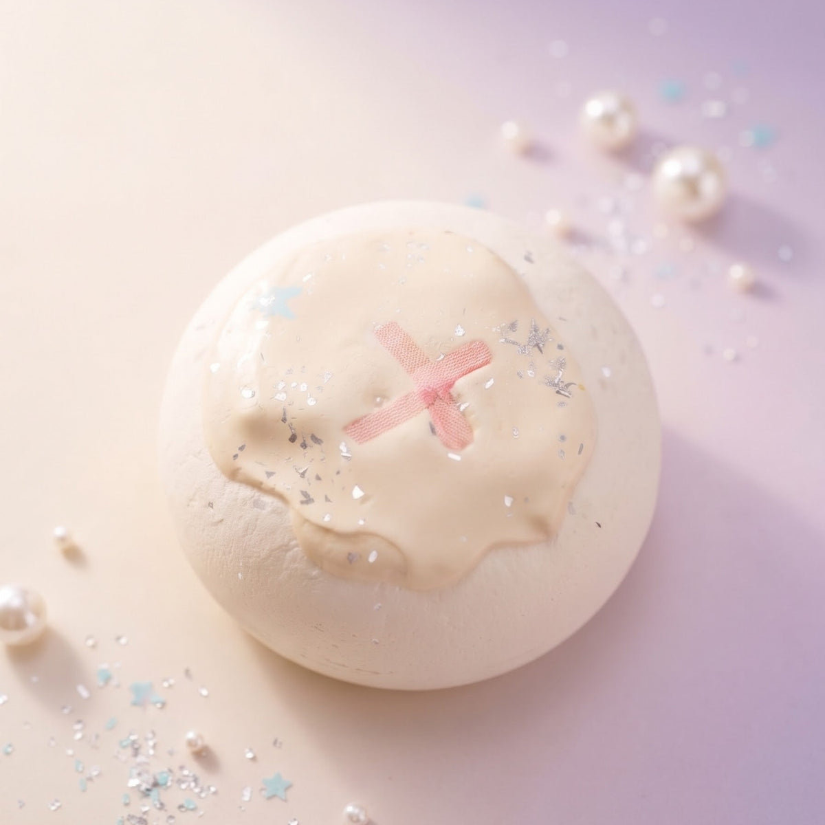 A round bath bomb with a glossy cream-colored surface, decorated with a pink 'X' and delicate blue star and snowflake details, resting on a pastel lavender background with scattered decorative pearls and glitter.