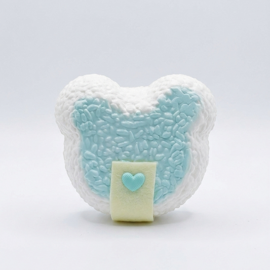 A heart-shaped bath bomb with a textured white exterior, a light blue textured center, and a pale yellow front flap featuring a small light blue heart, set against a seamless white background.