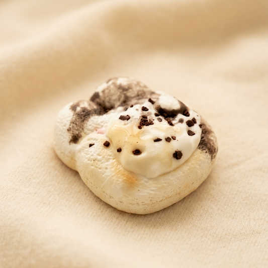 A small baked pastry or cookie with a white creamy topping and dark chocolate chips, resting on a soft beige fabric background.
