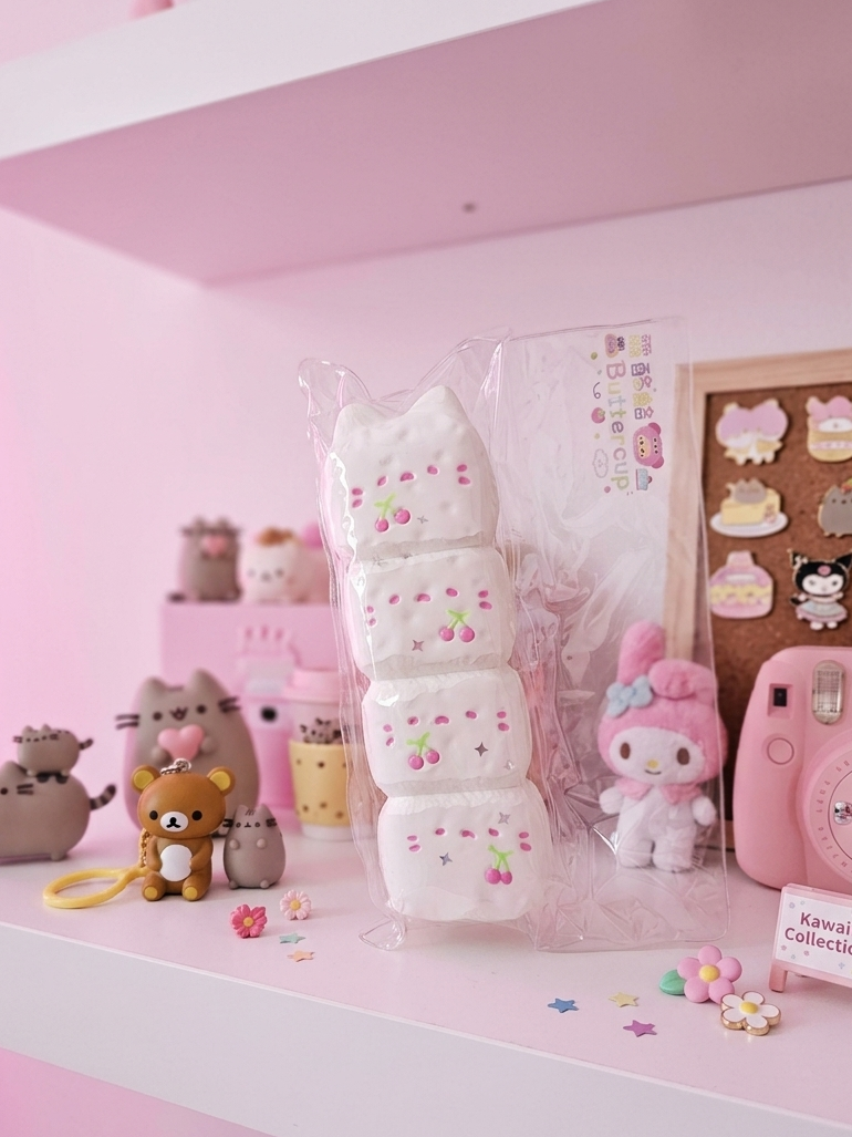 A pink-themed shelf display featuring a stack of three white marshmallow squishies with cherry details, a pink instant camera, plush toys, and cute accessories in a pastel pink room setting.