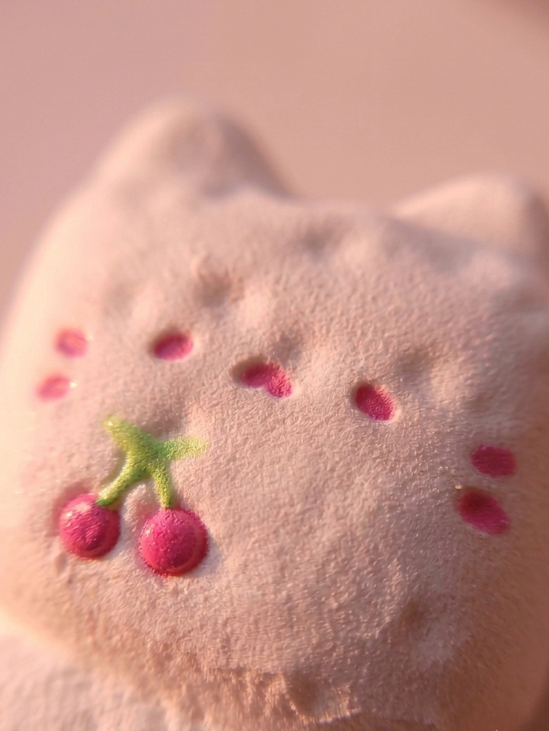 A close-up of a pale pink plush cat toy with embroidered pink cheeks, whiskers, and a small green and pink cherry motif on the front, set against a soft pink background.