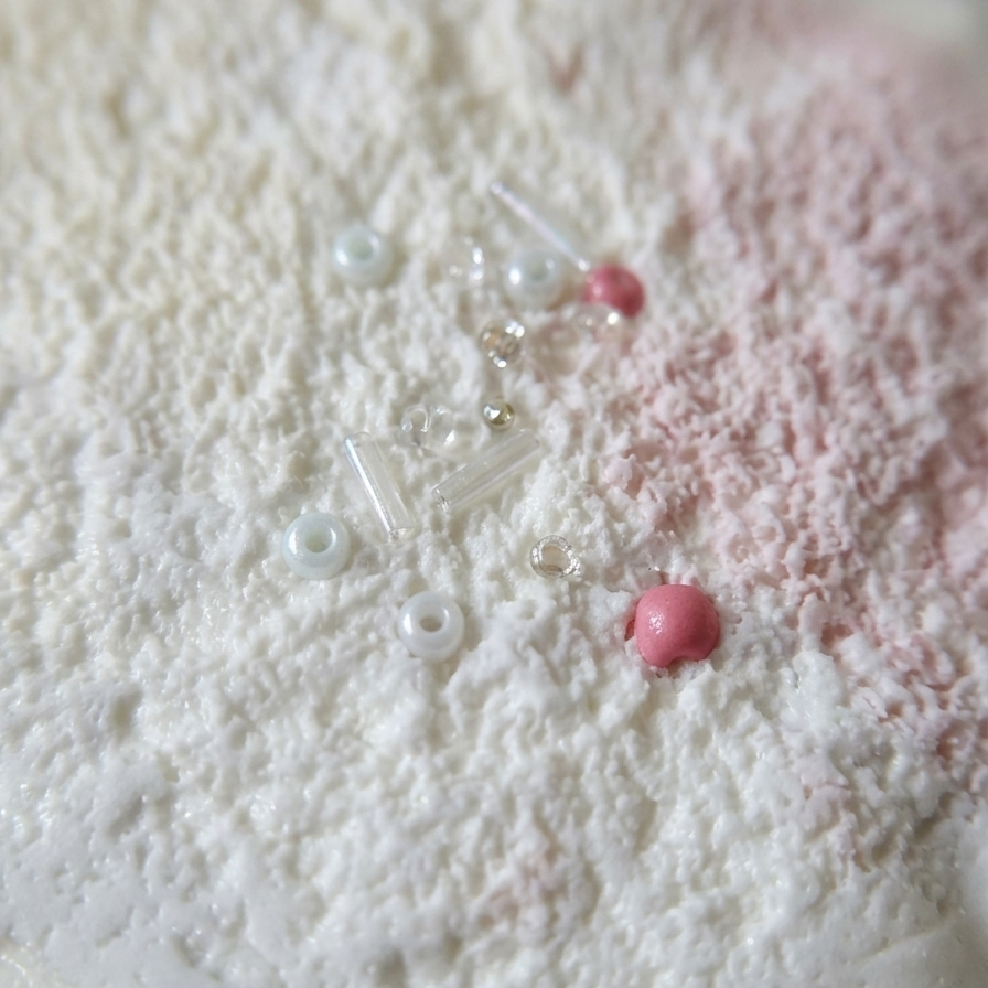 A close-up of a textured white surface with a light pink powder or dusting, accented by small clear beads, white pearl-like beads, and two pink round beads.