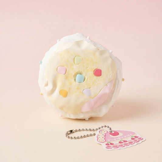A round, puffy white foam-like donut or macaron toy with pastel pink, yellow, blue, and pink sprinkles, shown on a soft pink background with a small matching keychain tag and ball chain in front.
