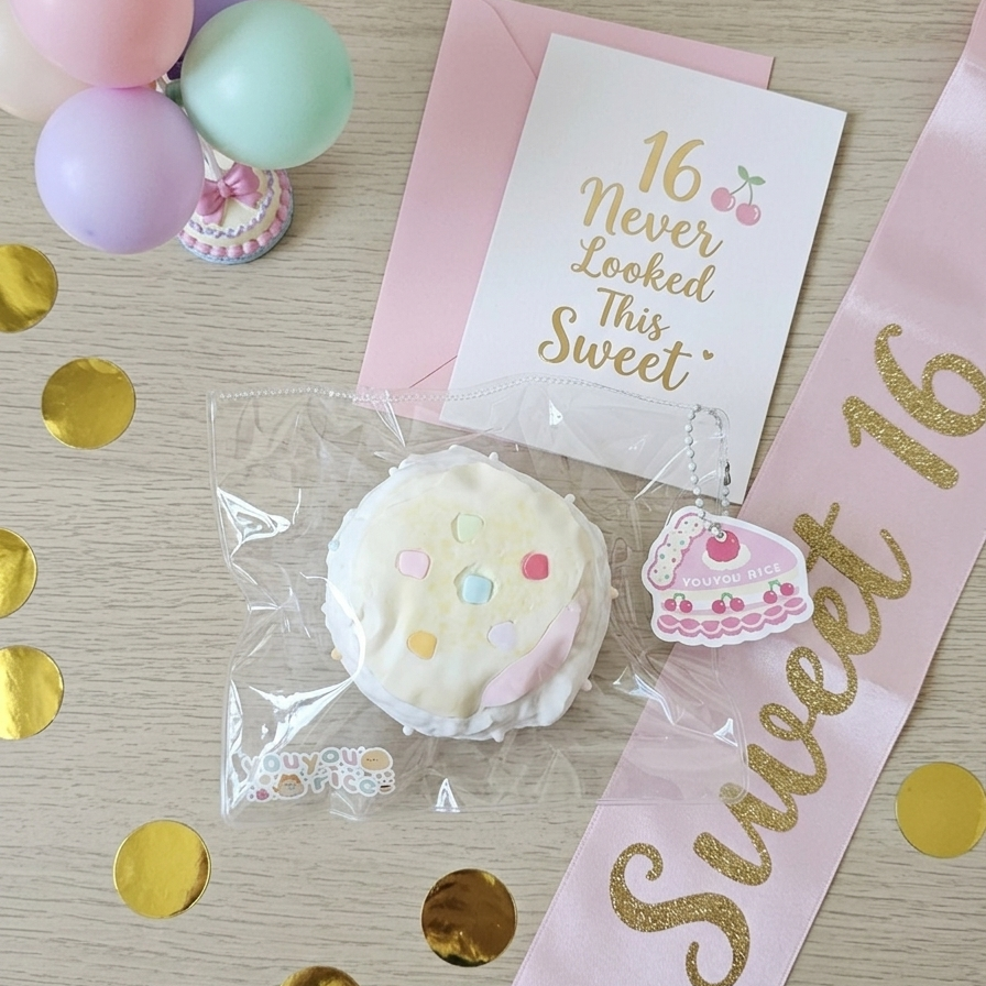 A styled flat-lay of a 16th birthday squishy toy set on a light wood surface with gold confetti, balloons, and a pink sash. The set includes a round frosted donut-like squishy with colorful sprinkles, a slice-of-cake charm, a pink envelope, a card reading '16 Never Looked This Sweet,' and a pink 'Sweet 16' sash.