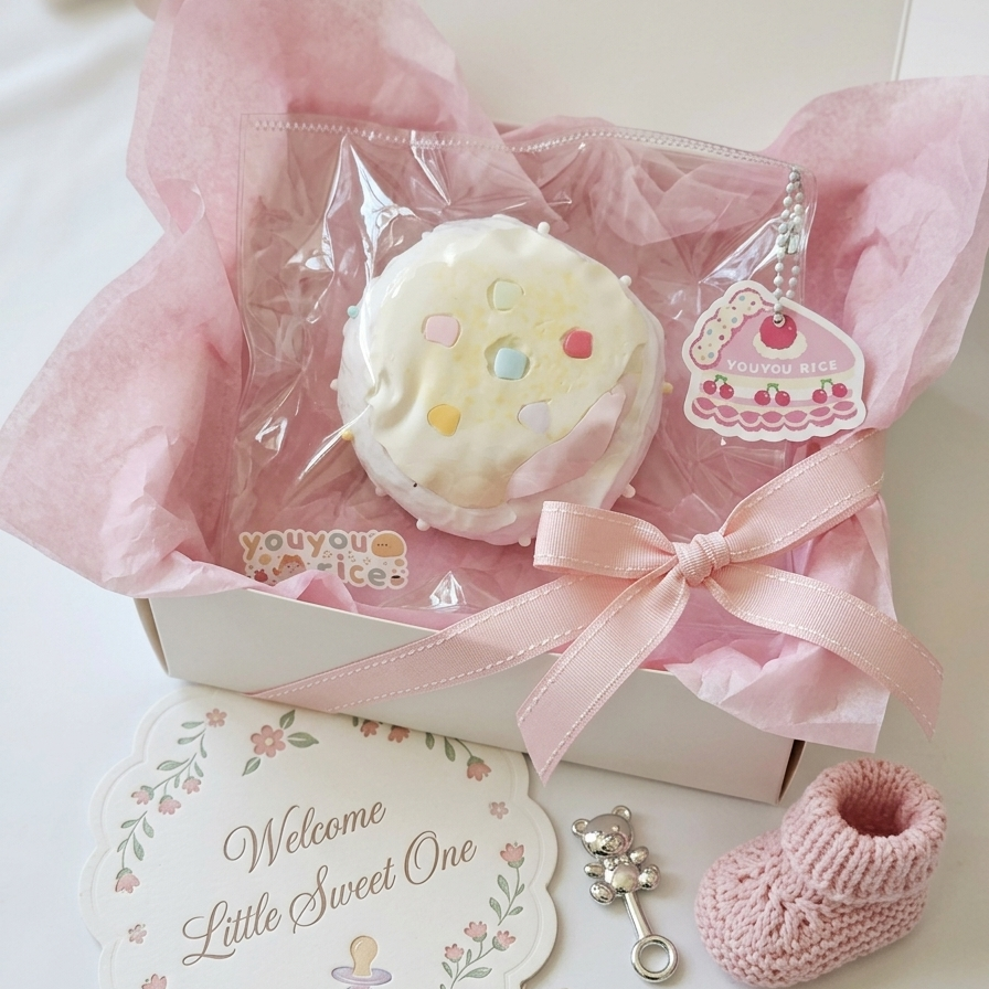 A baby shower gift box set featuring a white frosted donut-shaped plush toy with colorful sprinkles, a pink cake charm, a pink knit bootie, a 'Welcome Little Sweet One' card, and silver pacifier and key charms, all arranged in a white box with pink tissue paper.