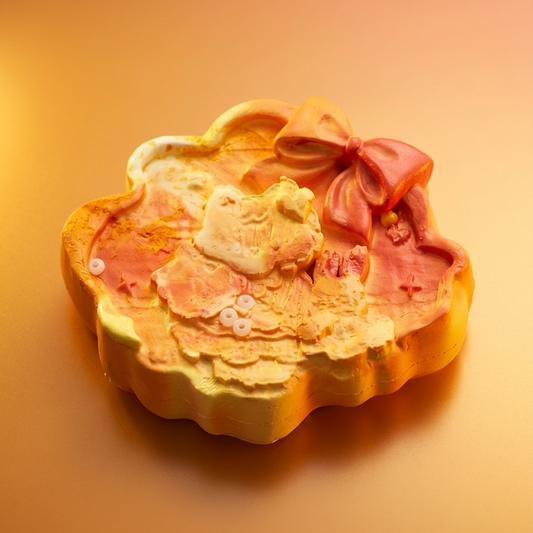 A decorative, cloud-shaped molded product in warm pastel tones, featuring a raised bow, layered floral texture, and a small Hello Kitty face motif, photographed on a smooth warm gradient background.