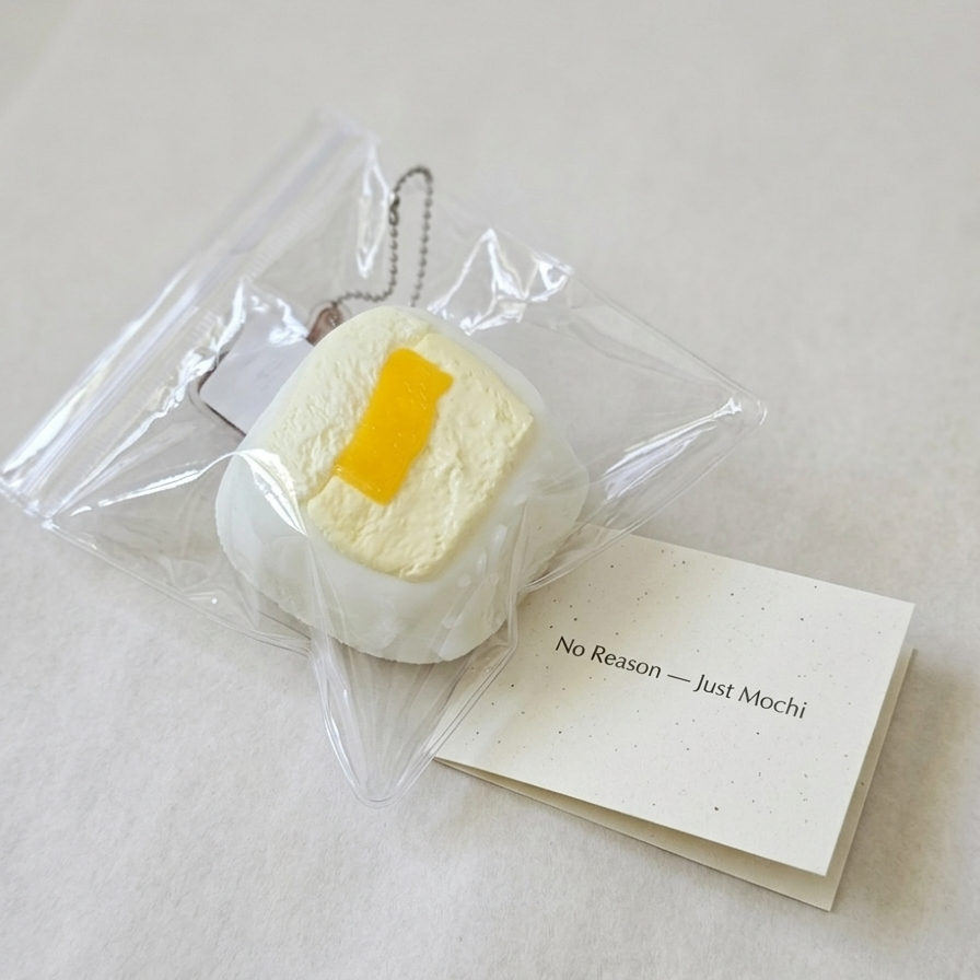 A small white mochi-shaped keychain or charm with a yellow butter-like accent, sealed in a clear plastic bag next to a cream card that reads 'No Reason - Just Mochi' on a light neutral background.