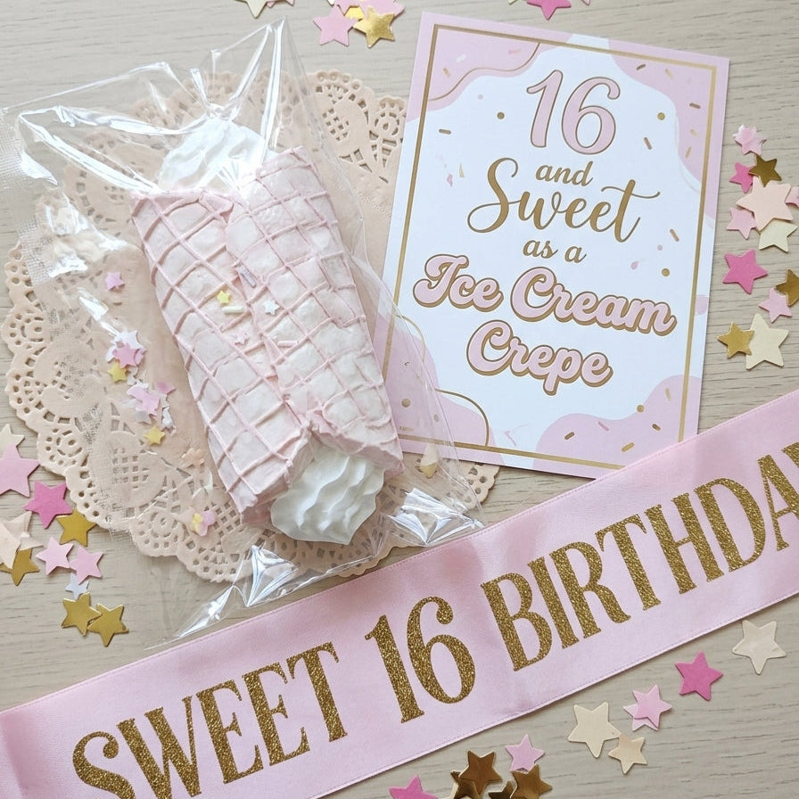 Pink and gold 16th birthday ice cream crepe themed party accessories, including a wrapped crepe, a '16 and Sweet as an Ice Cream Crepe' card, and a 'SWEET 16 BIRTHDAY' sash, arranged on a light wood background with star confetti.
