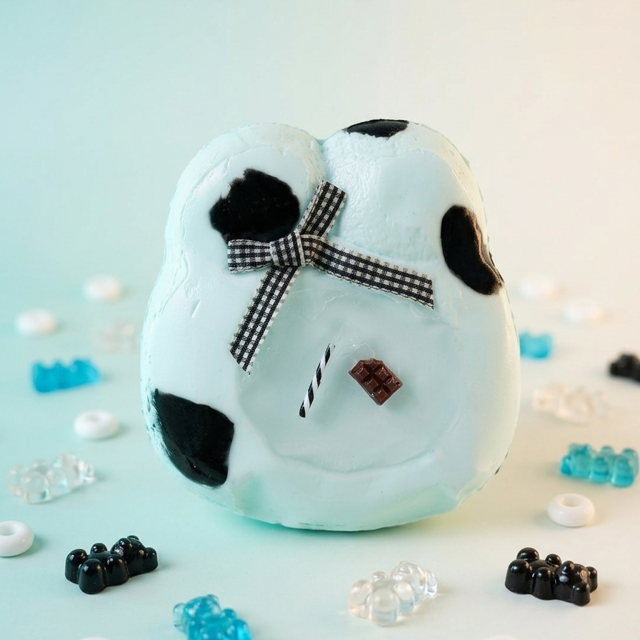 A white novelty toy or plush shaped like a cow with black spots, a black-and-white checkered bow, and small decorative facial details, photographed on a pale mint background with scattered candy-like props around it.