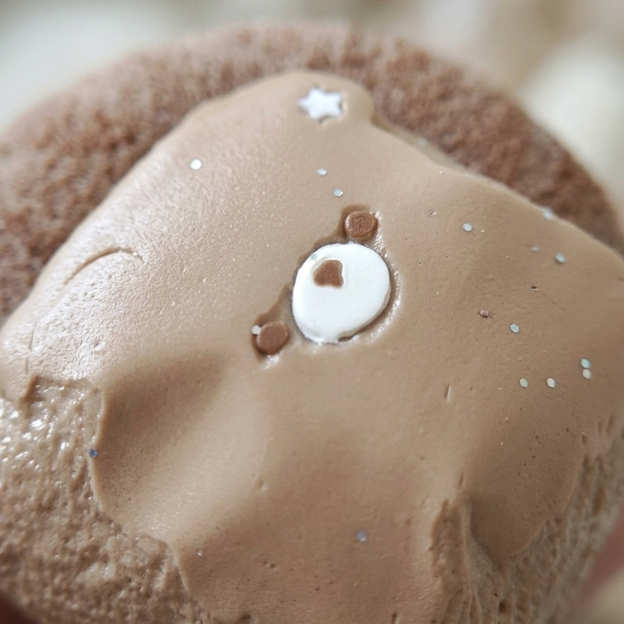 A close-up of a chocolate cake or pastry topped with smooth light brown frosting, decorated with a small white circular center, tiny brown dots, and scattered white sprinkles.