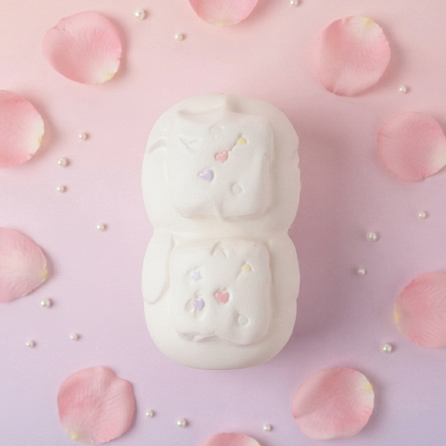 A white, double-bubble bath bomb shaped like a cute cartoon character, surrounded by scattered pink rose petals and small white pearls on a soft pink and purple gradient background.