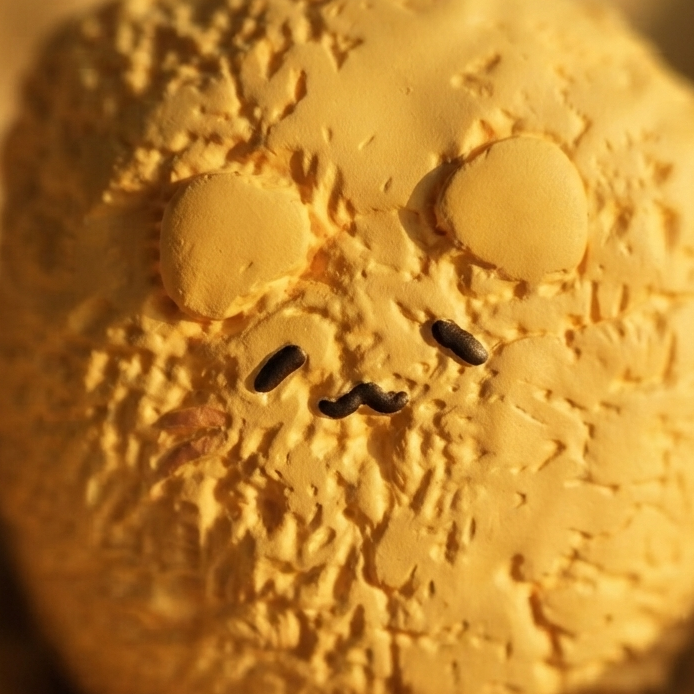 A close-up of a round, textured golden-yellow plush toy or decorative figure with simple embroidered facial features, resting on a soft beige fabric background.