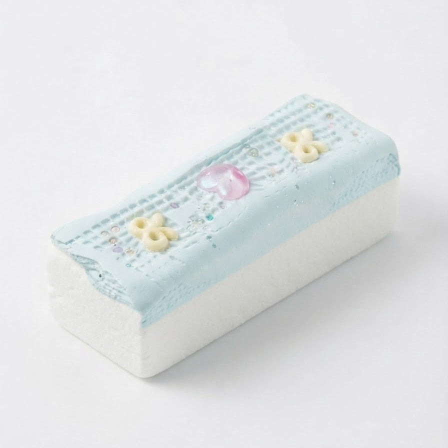 A rectangular decorative sponge or eraser with a light blue top featuring a grid pattern, embroidered details, a small pink heart, and yellow flower accents, resting on a white surface against a white background.