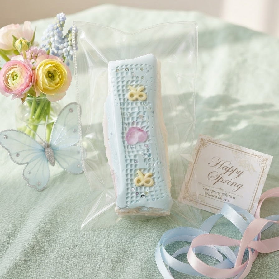 A packaged rectangular pastel blue item with embroidered floral details sits on a mint green fabric surface, styled with pink and yellow flowers, a butterfly decoration, a small card reading Happy Spring, and light blue and pink ribbons.