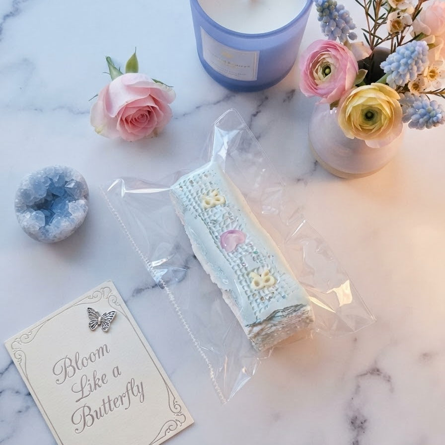 A packaged embroidered lace ribbon with pastel floral details lies on a white marble surface, styled with a blue candle, pink and yellow flowers, a small crystal, and a card that reads 'Bloom Like a Butterfly'.