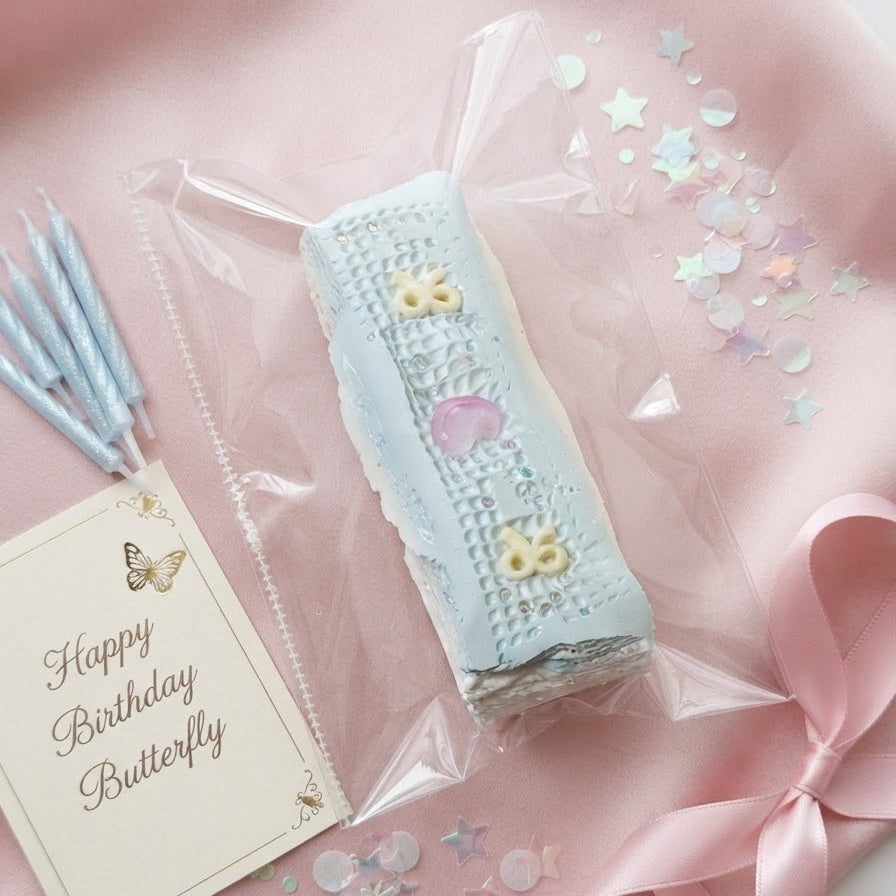 A wrapped pastel blue and pink birthday cake or pastry in clear cellophane packaging, styled on a pink fabric background with a pink ribbon, star confetti, blue candles, and a card that reads 'Happy Birthday Butterfly'.