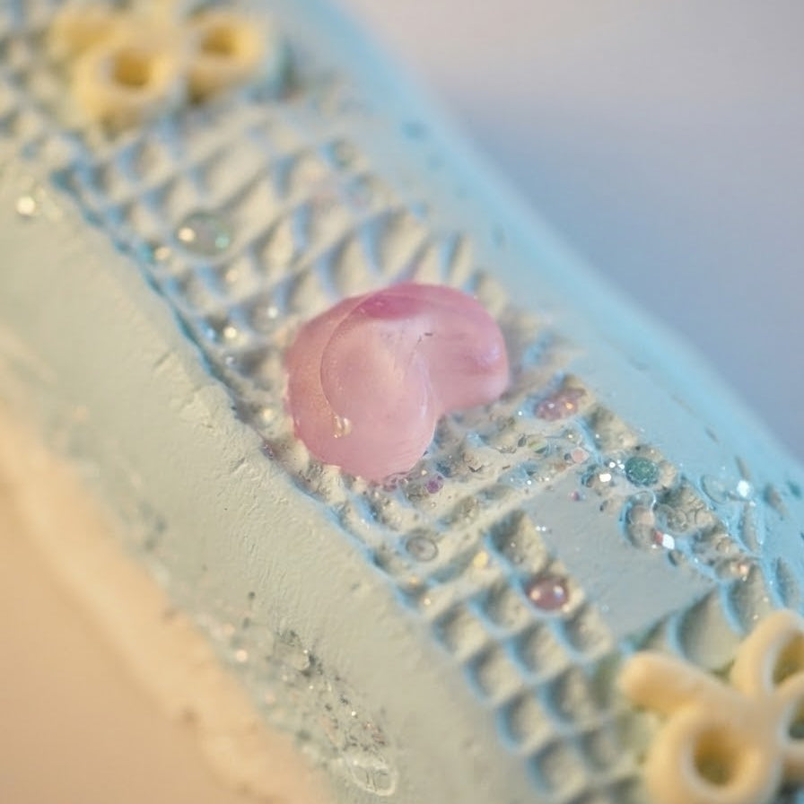 A close-up of a light blue textured decorative item with a small pink flower accent and pale yellow floral details, photographed against a soft neutral background.